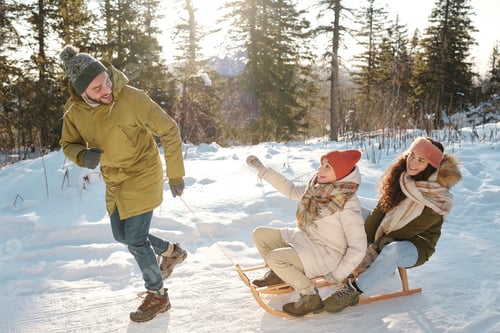 Preview: Two joyful girls in winterwear sitting on sledge while young man pulling them