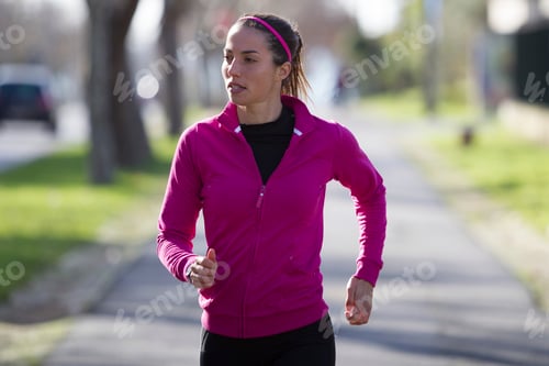 Preview: Focused female runner in a vibrant pink jacket jogging down a treelined path.