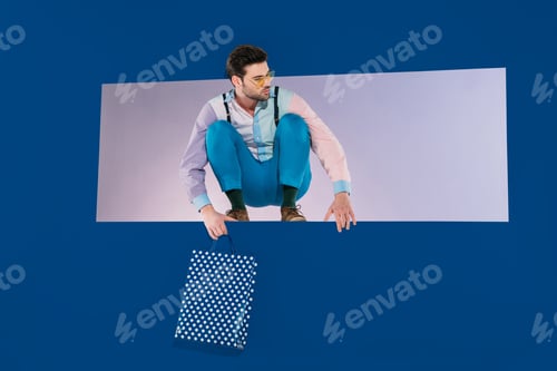 Preview: handsome man in stylish clothes squatting with shopping bag isolated on blue