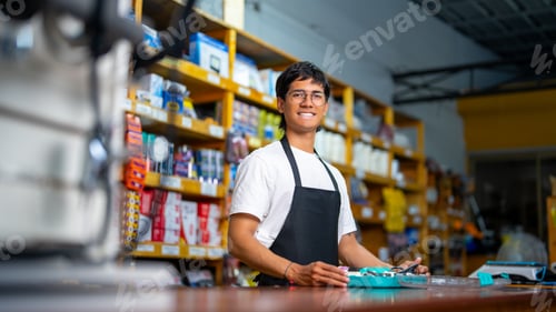 Preview: Young man wearing an apron, smiling while working at a retail store counter