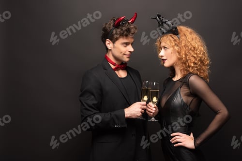 Preview: beautiful couple in halloween costumes clinking with champagne glasses on black