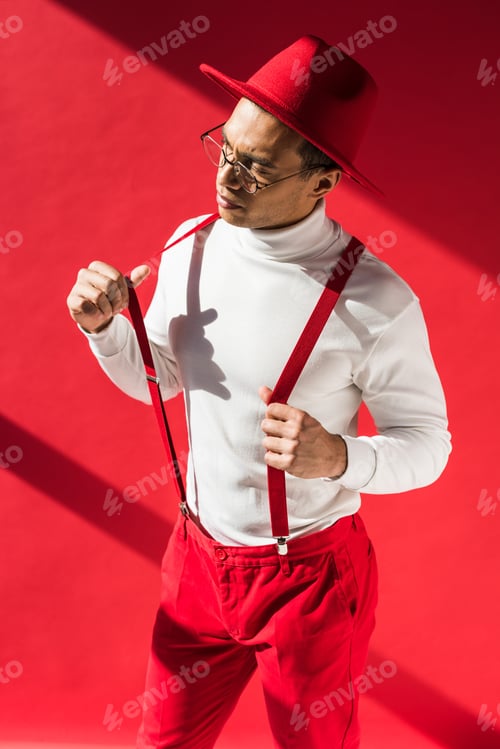 Preview: fashionable mixed race man in hat and suspenders posing on red
