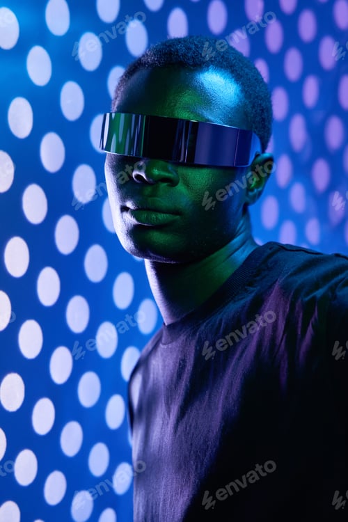 Preview: Young cool black in futuristic glasses standing near illuminated wall in studio