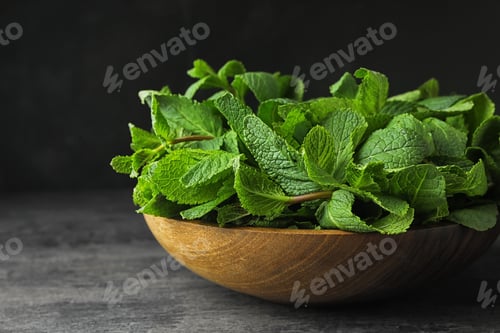 Preview: Wooden bowl full of fresh green mint on grey table against dark background, closeup. Space for text