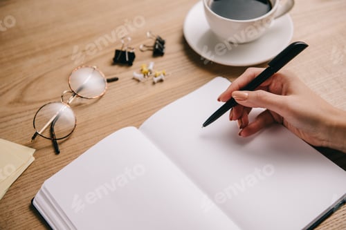 Preview: partial view of woman writing in notebook at workplace with eyeglasses and cup of coffee
