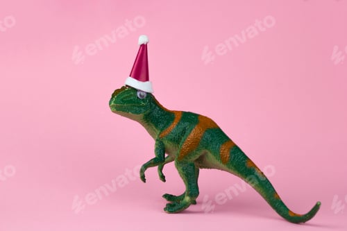 Preview: funny green dinosaur toy in little Santa Claus hat on pastel pink background