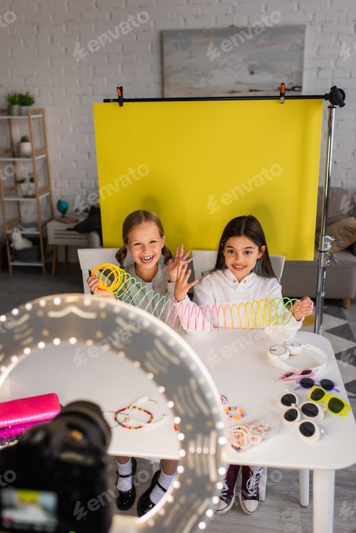 Preview: cheerful bloggers talking and showing spiral toy on yellow background in front of blurred digital