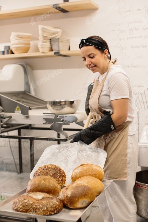 Preview: Female baker using a peel to take out a loaf of bread of the oven in a bakery