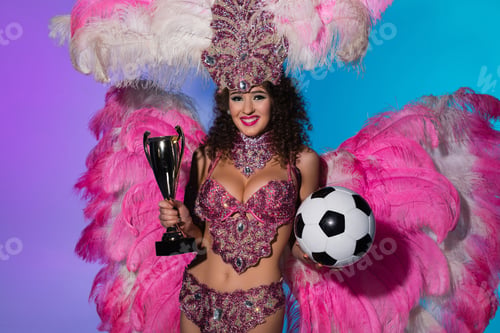Preview: Cheerful woman in carnival costume with pink feathers holding soccer ball and winner cup on blue