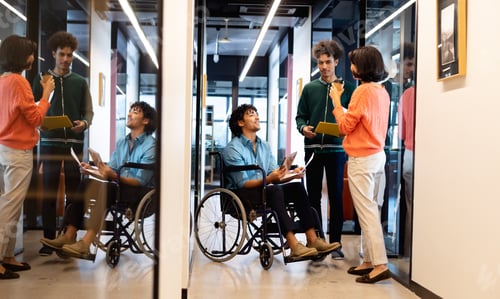 Preview: Multiracial colleagues discussing with young businessman sitting in wheelchair at office corridor