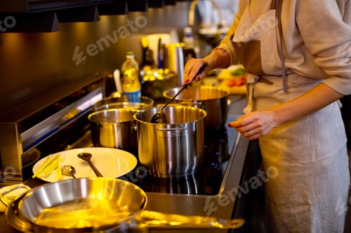 Preview: Woman in apron cooking pumpkin soup on induction oven on dark kitchen at home