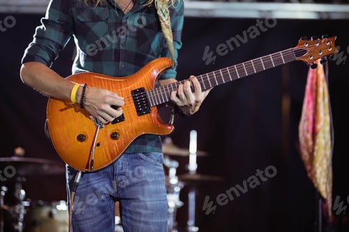 Preview: Male musician standing on stage, strumming orange guitar by drum kit and scarf-draped mic stand