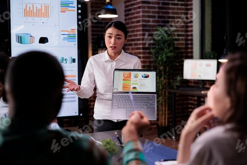 Preview: Asian woman in late night meeting holding laptop with business analytics discussing with coworkers