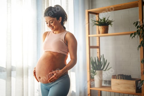 Preview: Fit pregnant woman holding hands on her belly, enjoying in the last trimester of pregnancy.