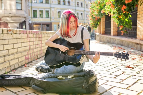 Preview: Creative female playing acoustic guitar sitting on sidewalk with guitar case and cash