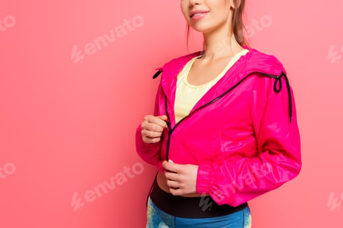 Preview: cropped view of young smiling sportswoman in pink windbreaker on pink background