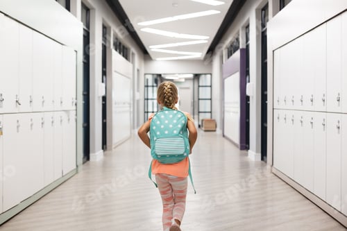 Preview: Rear view of elementary schoolgirl with backpack walking amidst lockers in corridor