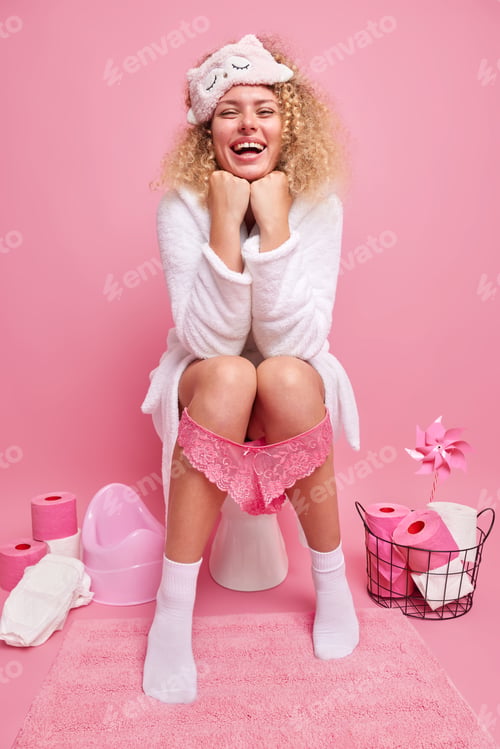 Preview: Indoor shot of happy curly haired young woman keeps hands under chin smiles broadly wears white bath