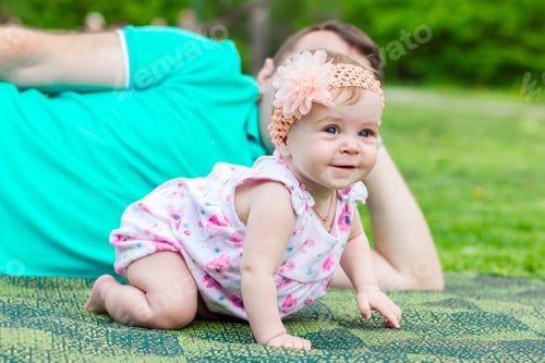 Preview: A funny baby with a headband with a flower is sitting with her dad on a green plaid in the garden.