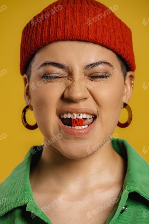 Preview: Playful young woman in hat holding plastic letters in mouth against yellow background