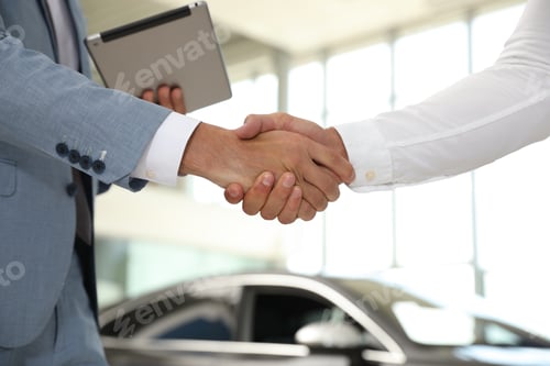 Preview: Young salesman shaking hands with client in modern car dealership, closeup