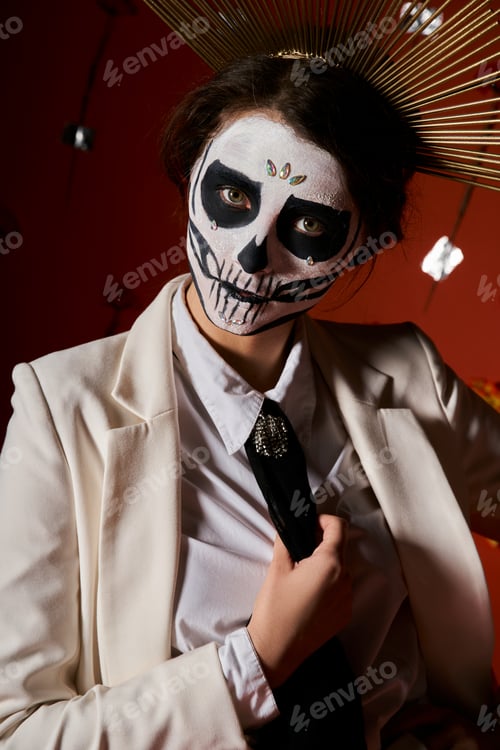 Preview: portrait of woman in skeleton makeup and white suit looking at camera on red backdrop, Day of Dead