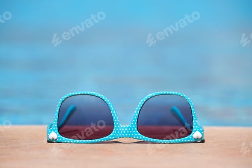 Preview: Closeup of blue sunglasses on swimming pool side on warm sunny day. Summer vacation concept.