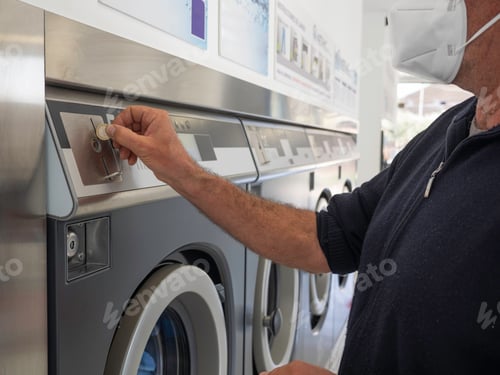 Preview: Man wearing a sanitary mask inserting a coin in a washing machine in a public laundromat