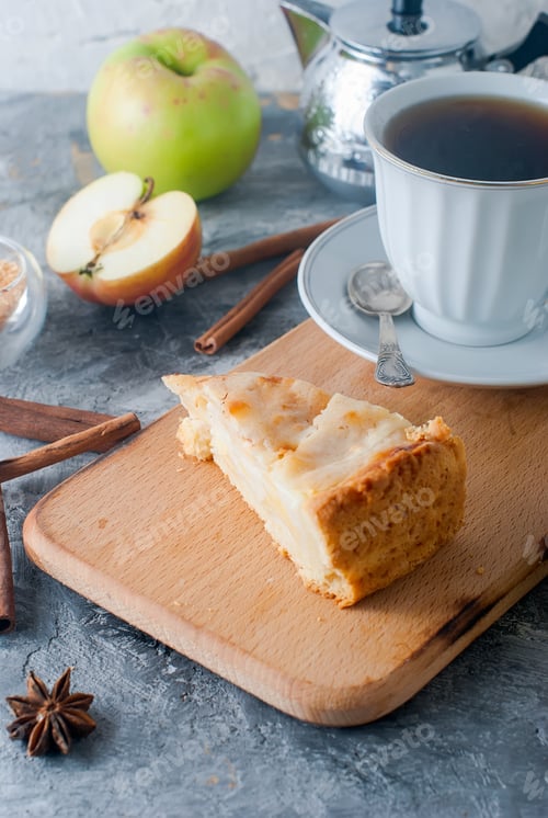 Preview: Apple pie sprinkled with icing sugar, cup of tea, apple and cinnamon sticks on table