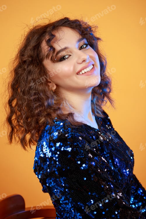 Preview: Portrait of young lady sittin on a chair on orange background and smiling to the camera