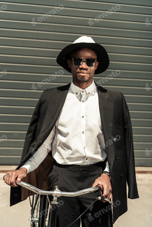 Preview: Fashionable african american businessman wearing fedora hat and holding bike