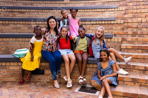 Preview: Portrait of diverse female teacher and schoolgirls sitting in elementary school outdoor auditorium