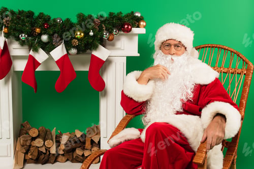 Preview: Santa claus sitting on rocking chair near fireplace with christmas stockings on green background