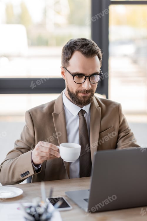 Preview: focused bearded businessman in stylish blazer, eyeglasses and tie holding coffee cup near mobile