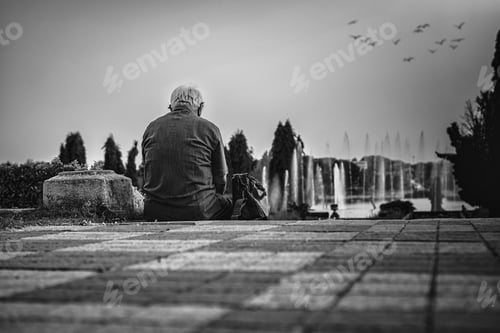 Preview: Beautiful shot of an old lonely man sitting on the ground and admiring a fountain