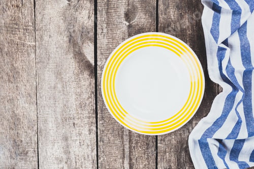 Preview: Empty white plate with yellow stripes and folded blue striped cloth on wooden table
