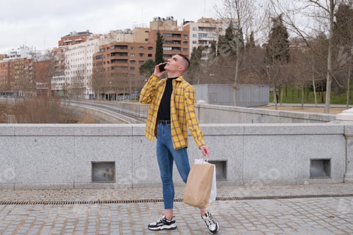 Preview: Young non binary person speaking on the smartphone with shopping bags. Make up androgynous person.
