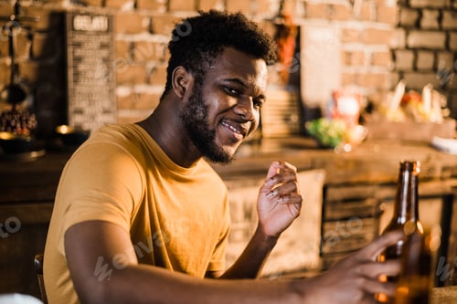 Preview: Young attractive black man with dark skin in the bar or kitchen home sitting with beer