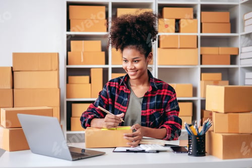Preview: A young woman is sitting at a desk with a laptop and a stack of boxes behind her