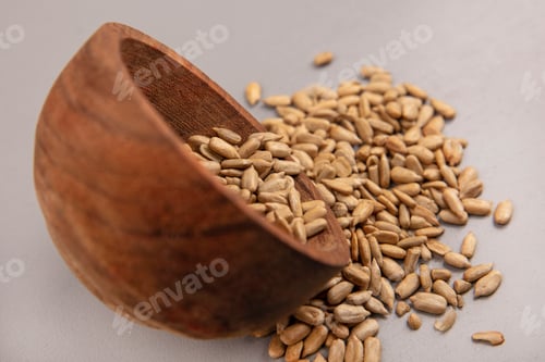 Preview: side view of organic shelled sunflower seeds falling out of a wooden bowl on a grey background