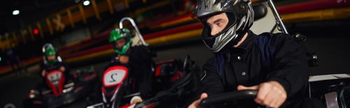Preview: concentrated man driving go kart near diverse drivers in helmets on indoor circuit, banner