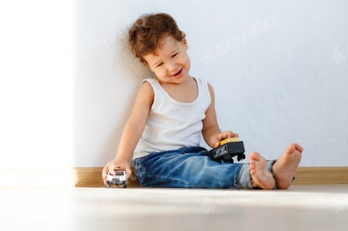 Preview: little boy on the floor plays cars dressed in white T-shirt and jeans, bright white room sunlight.