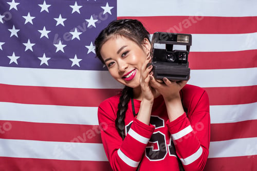 Preview: smiling asian woman holding retro photo camera with american flag behind