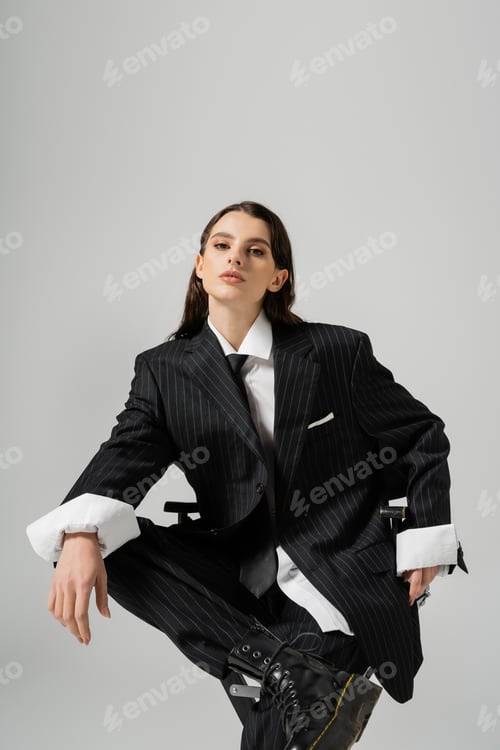 Vista previa: trendy woman in oversize formal wear sitting and looking at camera isolated on grey
