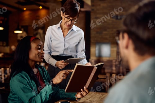 Preview: Happy waitress recommending order from a menu to young black woman in a bar.