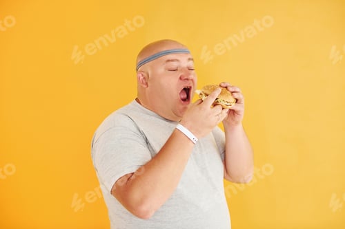 Preview: With fast food. Funny overweight man in sportive head tie is against yellow background