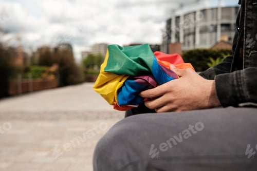 Preview: Young man sitting on a bench holding a crumpled rainbow flag