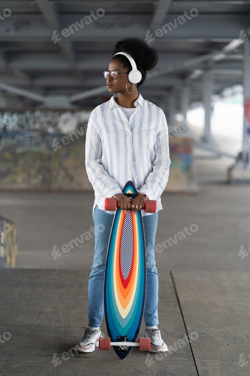 Preview: African girl with skateboard. Trendy female dressed in urban outfit wear headphones, hold longboard