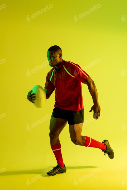 Preview: African american male rugby player running with rugby ball over yellow lighting
