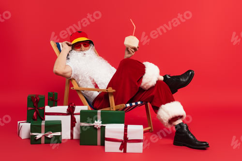 Preview: Santa claus in sunglasses holding cocktail in coconut on deck chair near gifts on red background